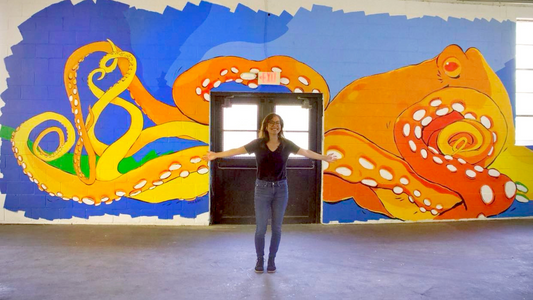 The artist posing in front of her big painted mural featuring an orange octopus and colorful ocean background.