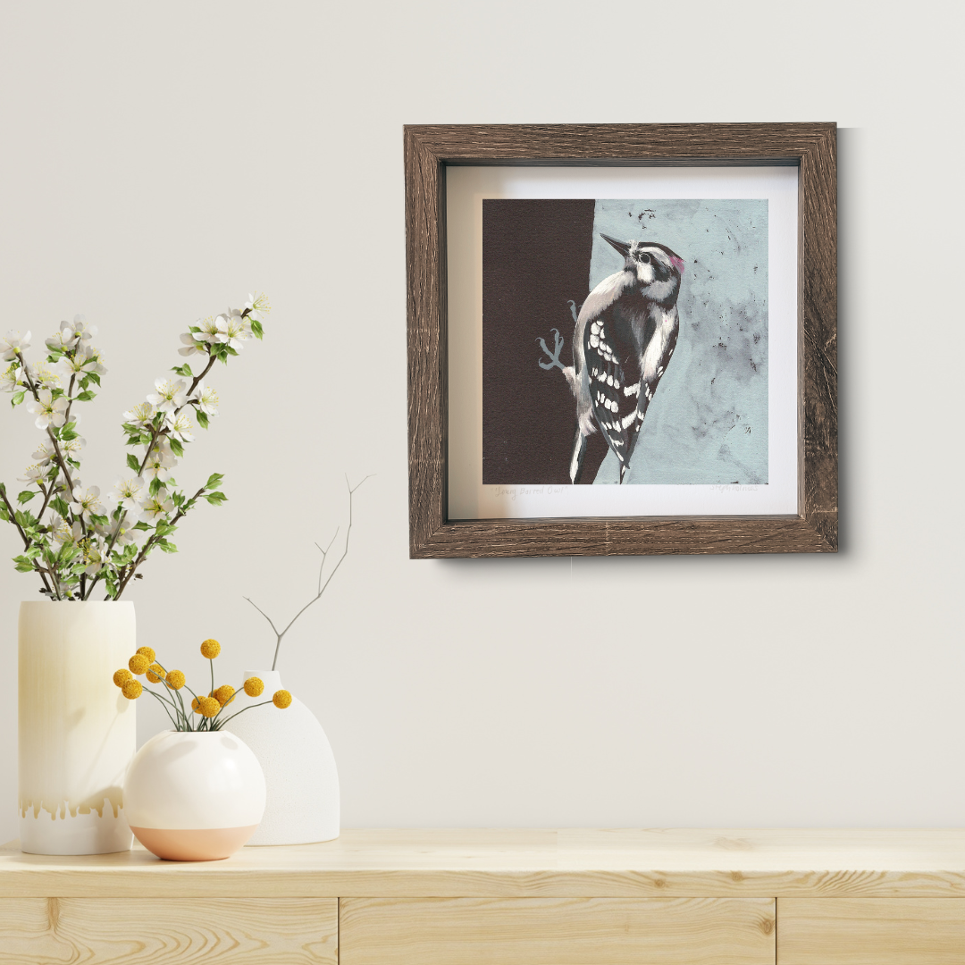 An art print featuring a Downy Woodpecker in a brown frame on a white wall above a modern wood desk.