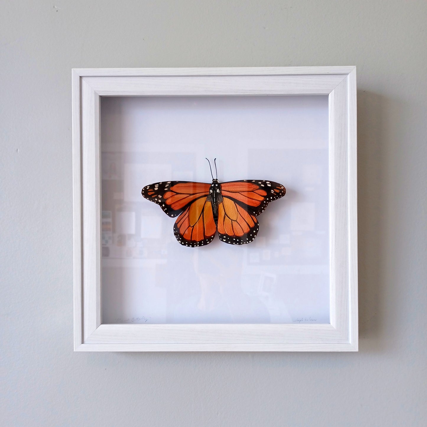 An orange monarch butterfly sculpture sits mounted in a white shadowbox.