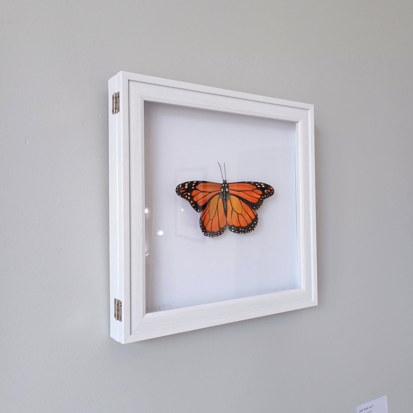 An orange monarch butterfly sculpture sits mounted in a white shadowbox.