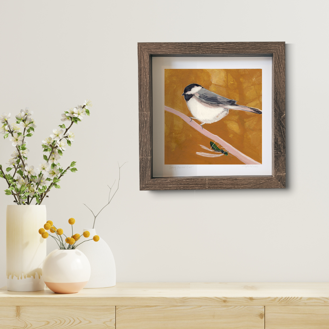 An art print featuring a black capped chickadee bird with a yellow background and a little green wasp. It's in a brown frame on a white wall above a modern wood desk.