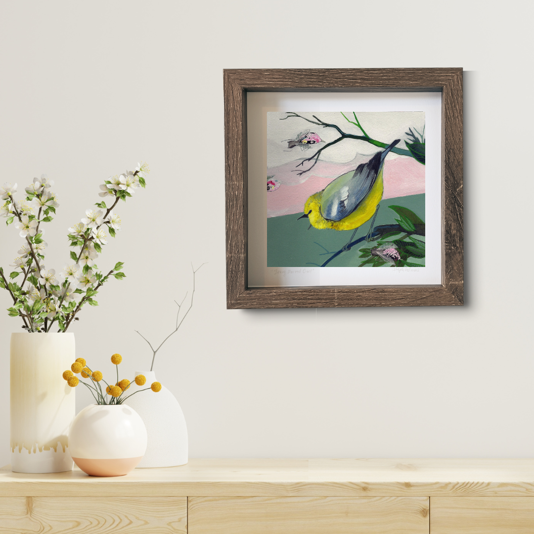 An art print featuring a bright blue winged warbler bird in a brown frame hanging above a modern wood desk.