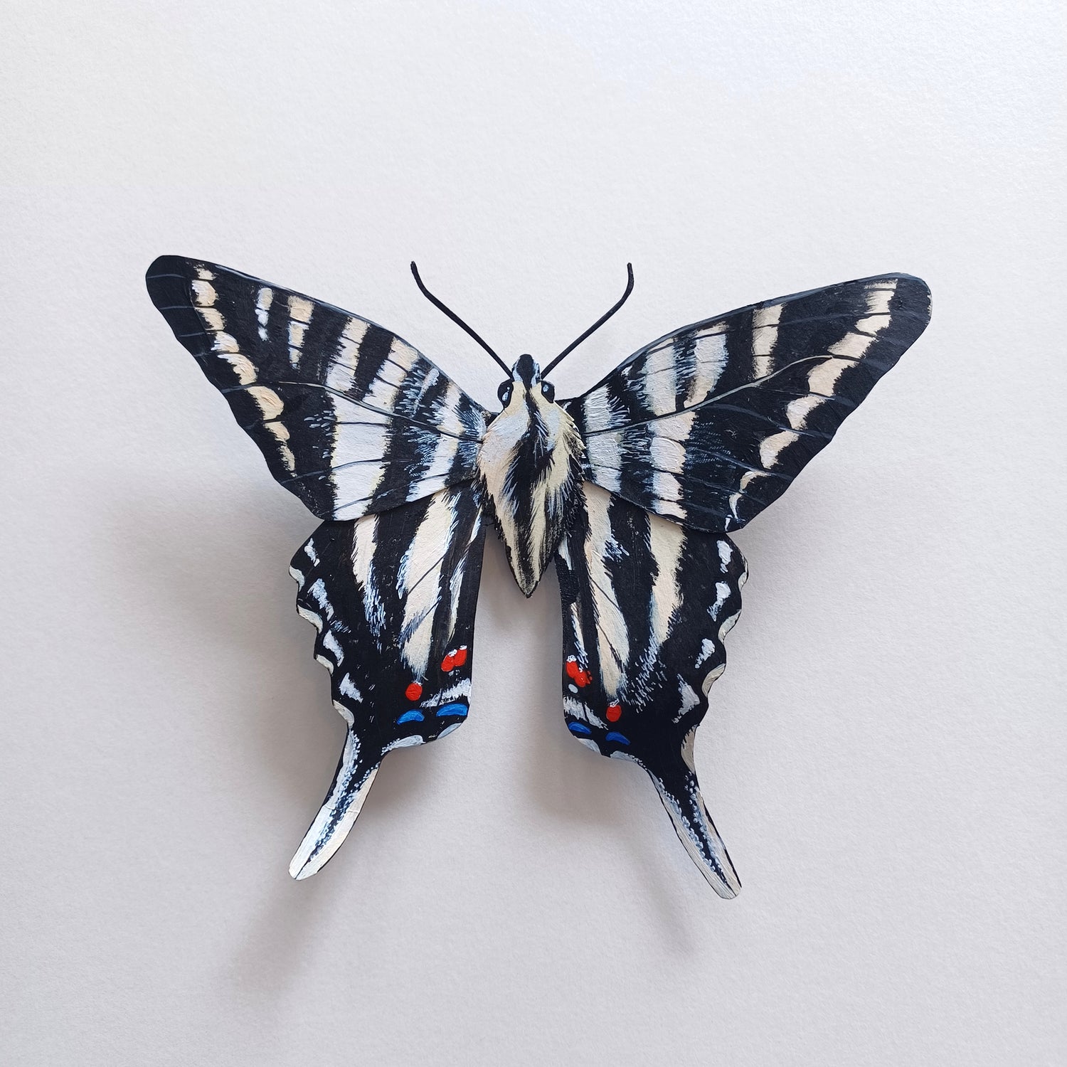 A photo of a painted paper sculpture of a zebra swallowtail butterfly in a white shadowbox.