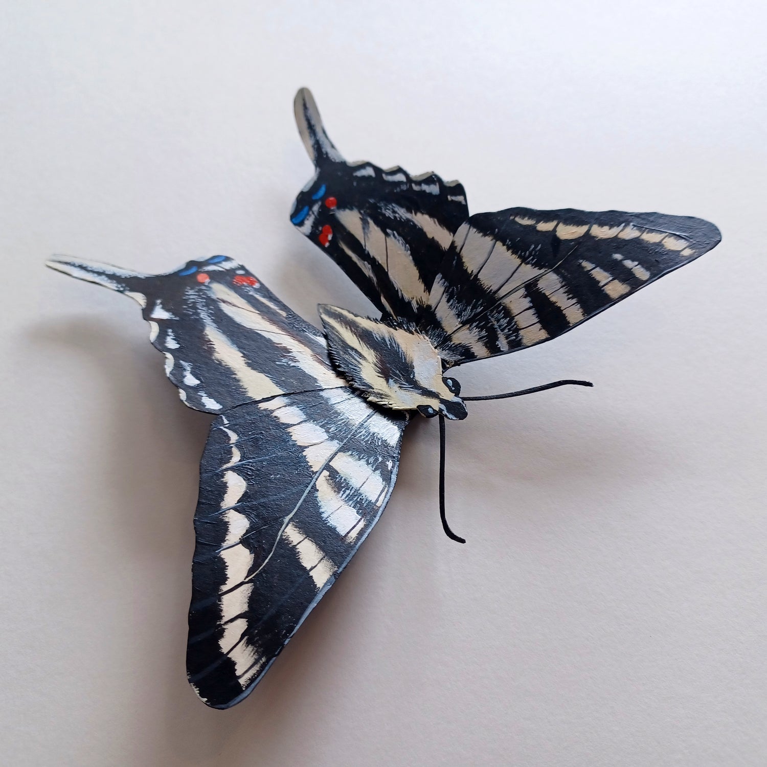 A photo of a painted paper sculpture of a zebra swallowtail butterfly in a white shadowbox.