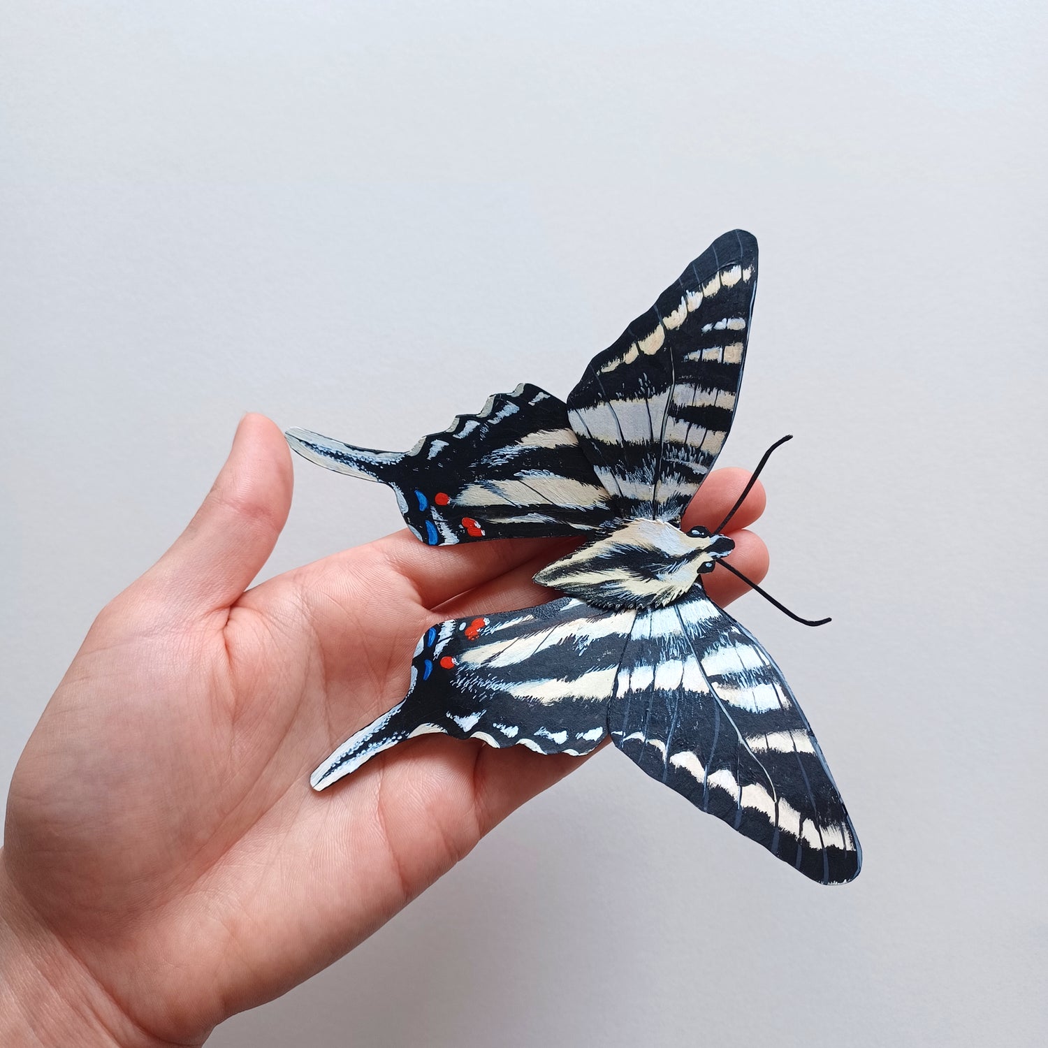 A photo of a painted paper sculpture of a zebra swallowtail butterfly in a white shadowbox.